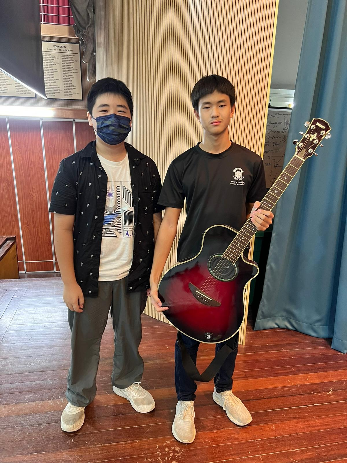 Two young men standing indoors, one wearing a face mask, the other holding a red and black acoustic guitar.